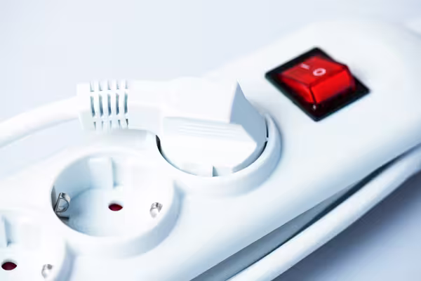 Nahaufnahme einer weißen Mehrfachsteckdose. Ein weißer Stecker ist in eine der Steckdosenbuchsen eingesteckt. Rechts daneben befindet sich ein roter Kippschalter mit der Ein-/Aus-Markierung „I/O“. Die restlichen Steckplätze sind leer. Der Hintergrund ist hell und schlicht, wodurch der Fokus klar auf der Steckdose und dem eingesteckten Stecker liegt.