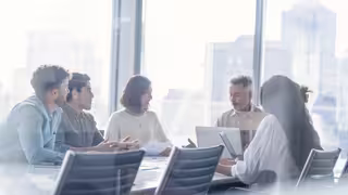 Foto: Ein fünfköpfiges Team sitzt an einem Tisch vor einer Fensterfront und tauscht sich aus.