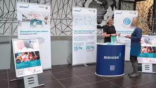Aktionswoche im Landeskriminalamt NRW. Eine Informations- und Beratungsstation der Kampagne „mission E“ in einem Foyer. Mehrere Roll-up-Banner mit Texten und Bildern stehen hinter einem runden, blauen Infostand, der das Kampagnenlogo trägt. Zwei Frauen stehen am Stand und sprechen miteinander.