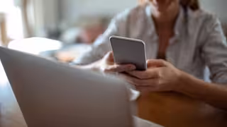 Eine Person sitzt an einem Tisch und hält ein Smartphone in beiden Händen. Vor ihr steht ein geöffneter Laptop. Der Hintergrund ist unscharf, die Szene wirkt wie ein Homeoffice- oder Arbeitsumfeld. Die Person trägt ein helles Oberteil, ihr Gesicht ist nicht vollständig zu sehen.