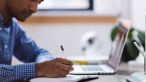 Mann sitzt am Schreibtisch vor seinem Laptop und schreibt mit einem Stift in ein Buch.