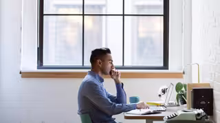 Person sitzt an einem Schreibtisch vor einem Laptop und arbeitet konzentriert in einem hellen Raum mit großem Fenster.