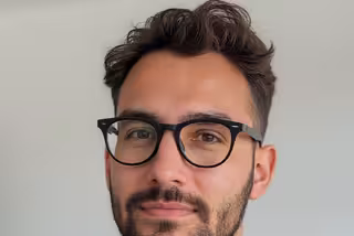 Herr Müller trägt eine Brille. Er hat kurze braune Haare und einen Dreitagebart.