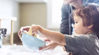 Kind und erwachsene Person spülen gemeinsam Geschirr an einem Waschbecken, beide halten eine schaumige Schüssel.