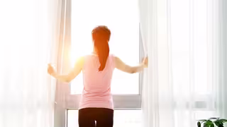 Eine Person steht in einem hellen Raum vor einem großen Fenster. Sie hält mit beiden Händen die weißen Vorhänge zur Seite und blickt nach draußen ins Sonnenlicht. Die Szene wirkt ruhig und frisch, das Licht fällt sanft in den Raum. Links und rechts des Fensters hängen leichte, helle Gardinen. Rechts im Bild ist eine grüne Zimmerpflanze zu sehen.