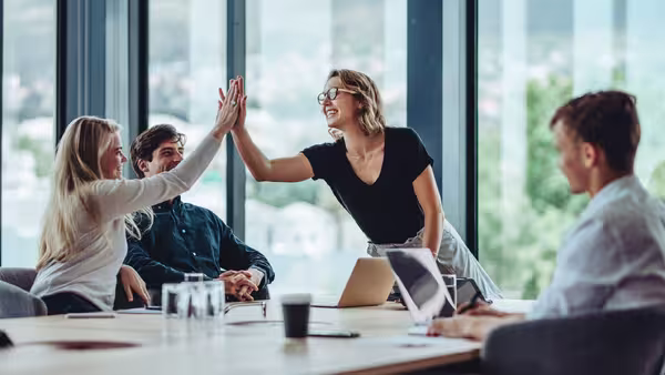 Foto: vier gutgelaunte Menschen an einem Besprechungstisch, zwei Frauen klatschen sich ab